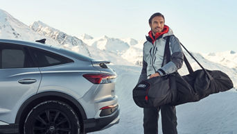 Mann in Winterkleidung trägt Audi-Skitasche neben silbernem Audi vor verschneiter Bergkulisse.