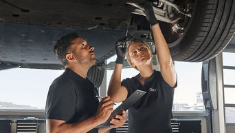 Zwei Audi-Mitarbeitende inspizieren gemeinsam die Fahrzeugunterseite in einer hellen Werkstattumgebung.